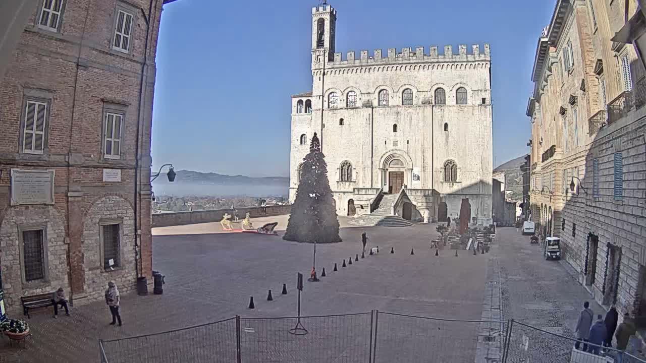 A grand historic stone building with a bell tower and battlements dominates a town square on an overcast day, featuring a large undecorated evergreen tree, various vehicles, scattered people, and foreground construction work, all set against distant hills.