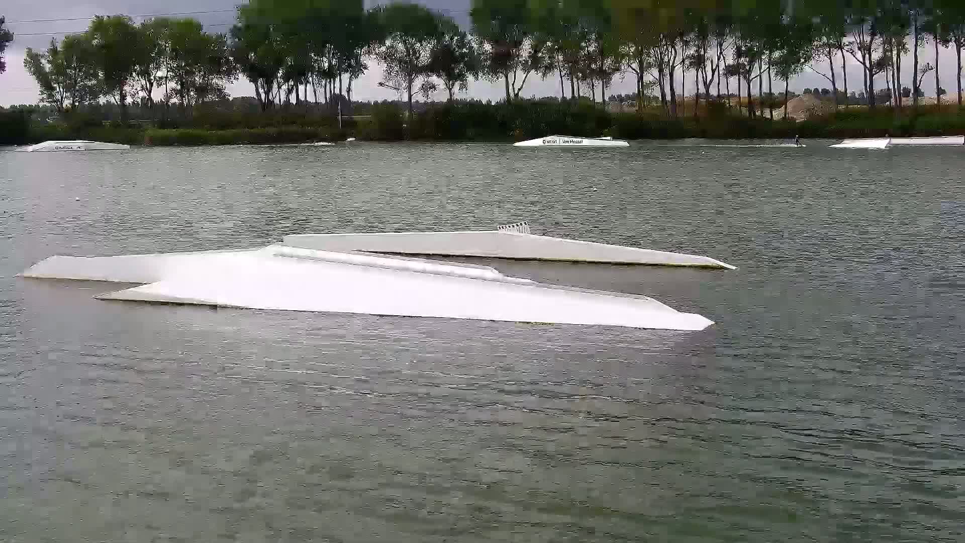 Several white ramps for watersports are partially submerged in a calm body of water under an overcast sky, surrounded by trees.
