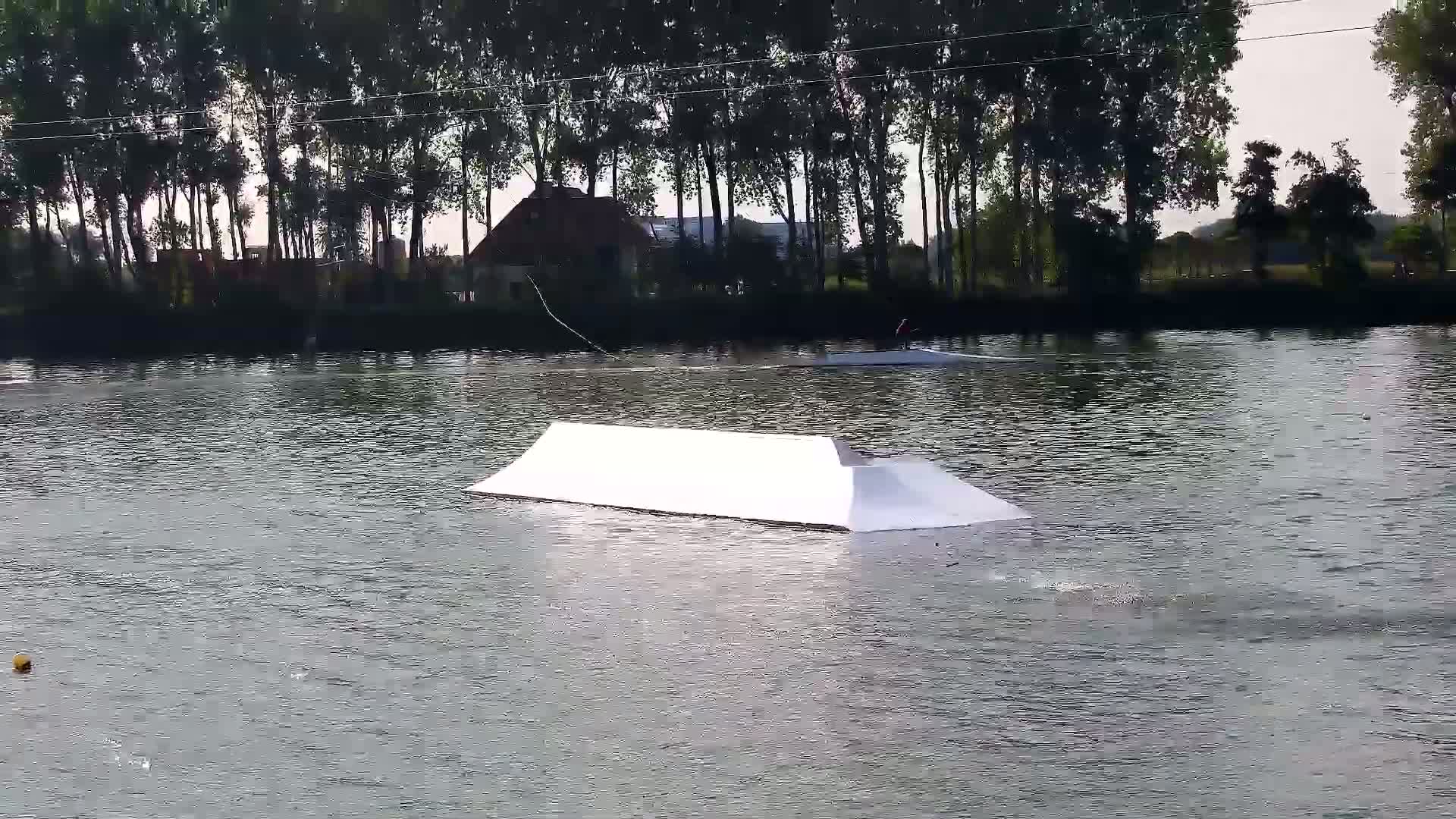 A white wakeboarding ramp floats in a calm body of water bordered by a line of trees under an overcast sky.