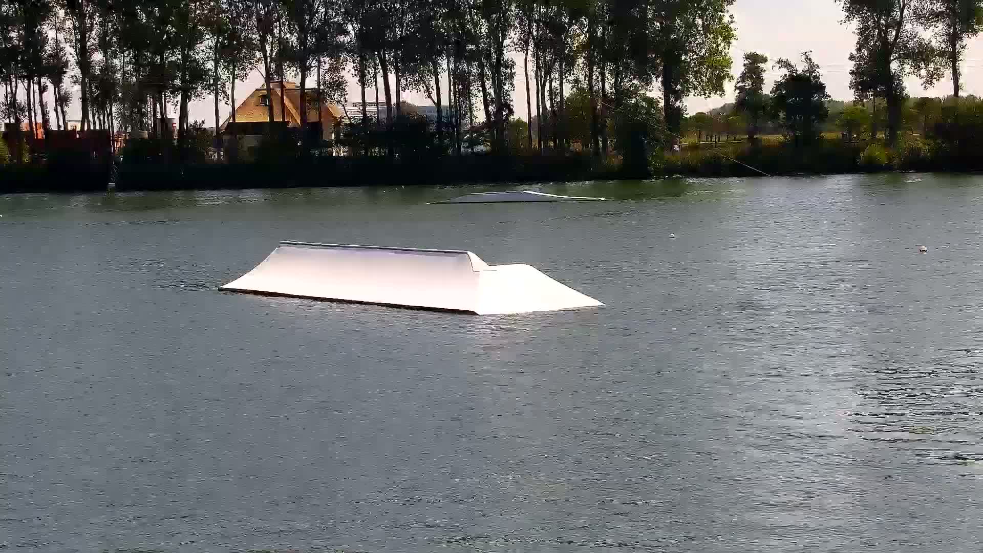 A white wakeboarding ramp floats in a calm body of water bordered by a line of trees under an overcast sky.