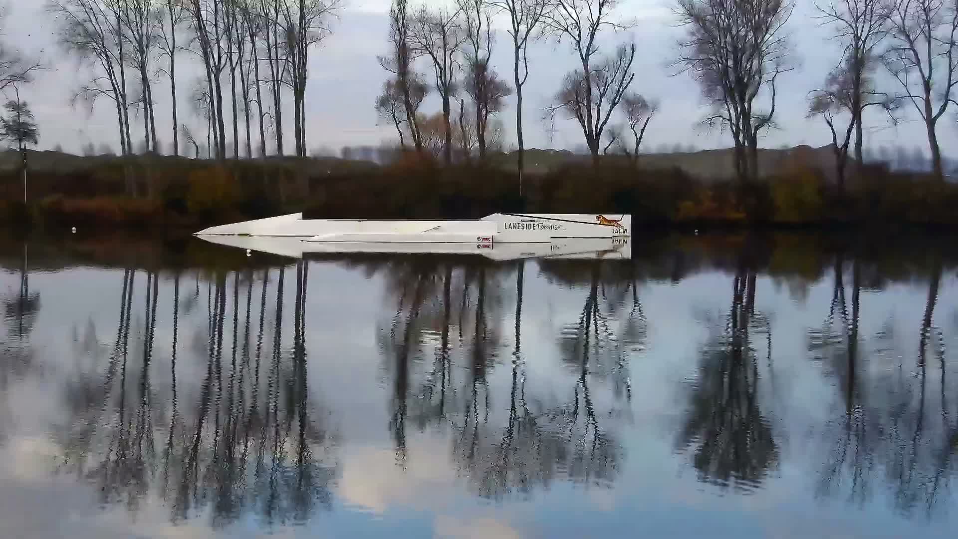 A long white boat rests on calm, reflective water mirroring a line of bare trees along a distant shore under an overcast sky, indicating a cool, tranquil day.