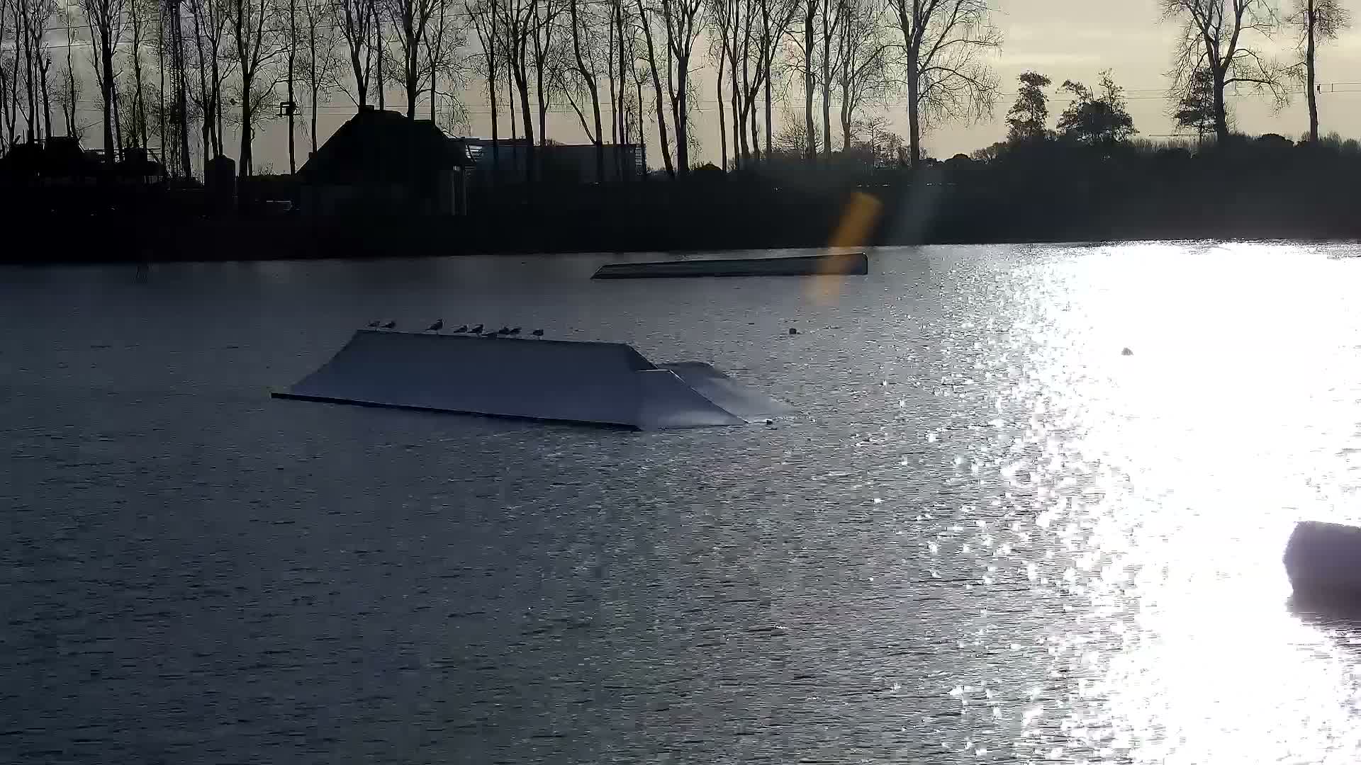 A long white boat rests on calm, reflective water mirroring a line of bare trees along a distant shore under an overcast sky, indicating a cool, tranquil day.