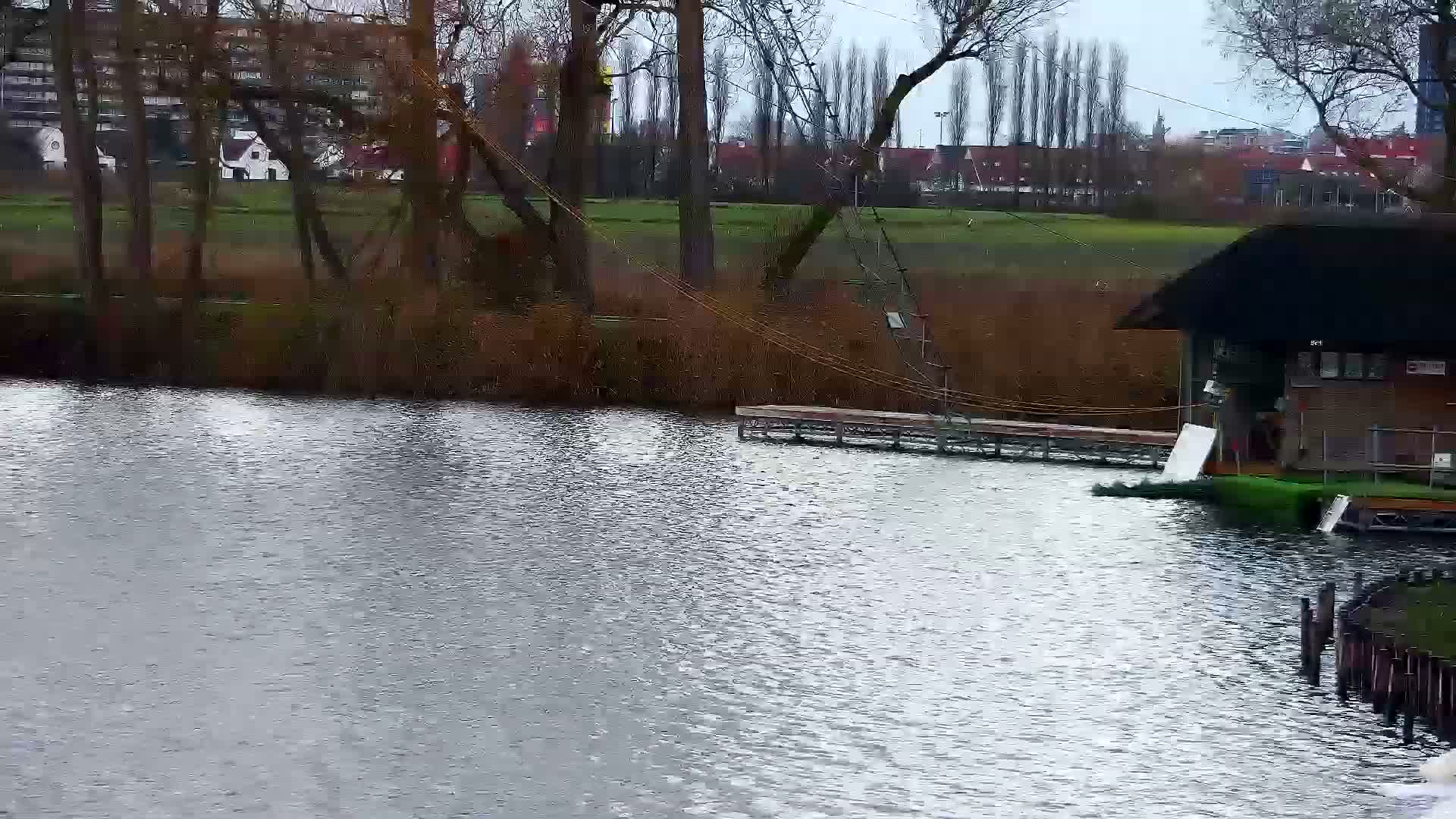 An overcast sky hangs over a rippling body of water in the foreground, leading to a grassy bank adorned with bare trees, a boathouse, a wooden dock, and a cable-skiing lift system, with distant buildings peeking through the foliage.