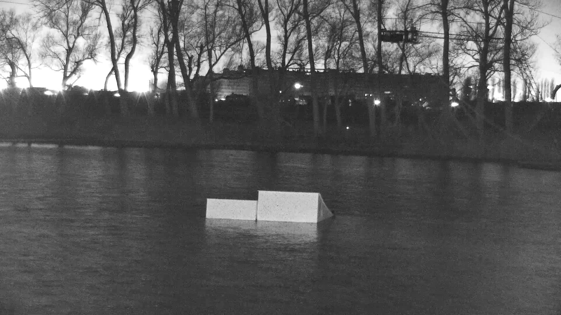 A desaturated, nighttime image features two geometric obstacles floating on a rippling body of water, backed by a dark line of bare trees and distant urban lights under a chilly, overcast sky.