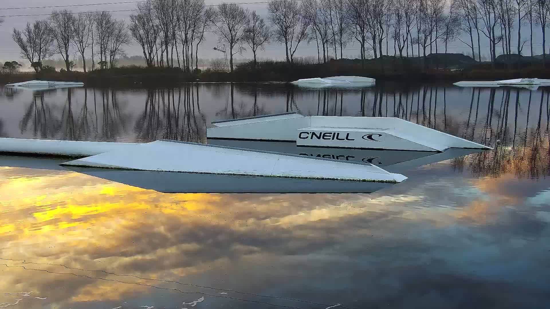 White wakeboard obstacles float on calm water reflecting a vibrant mix of golden, orange, and gray clouds, with a line of bare trees on the distant shore under an overcast sky.