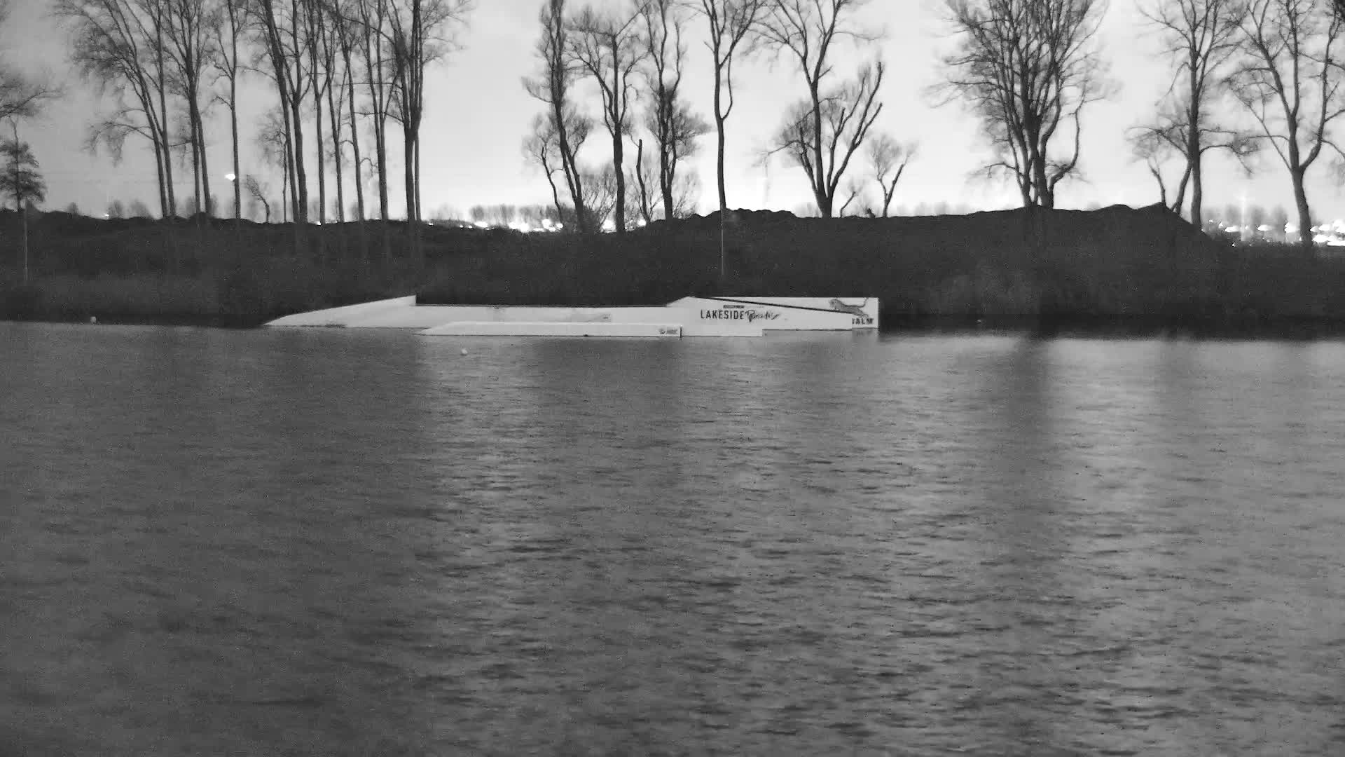 White wakeboard obstacles float on calm water reflecting a vibrant mix of golden, orange, and gray clouds, with a line of bare trees on the distant shore under an overcast sky.