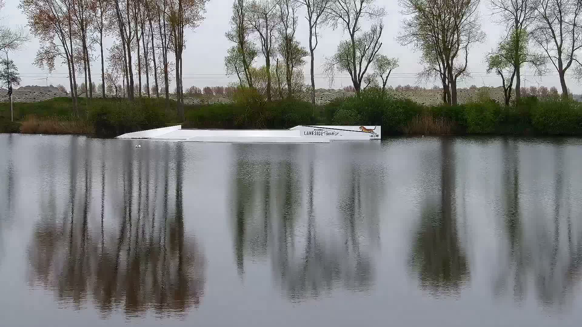 White wakeboard obstacles float on calm water reflecting a vibrant mix of golden, orange, and gray clouds, with a line of bare trees on the distant shore under an overcast sky.