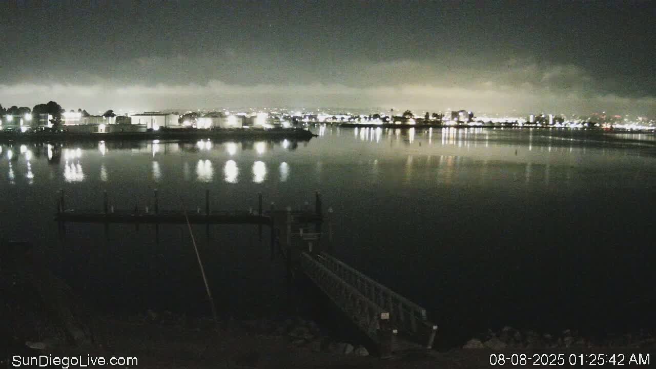 A nighttime view of a calm body of water reflecting the lights of a city skyline under a slightly cloudy sky.