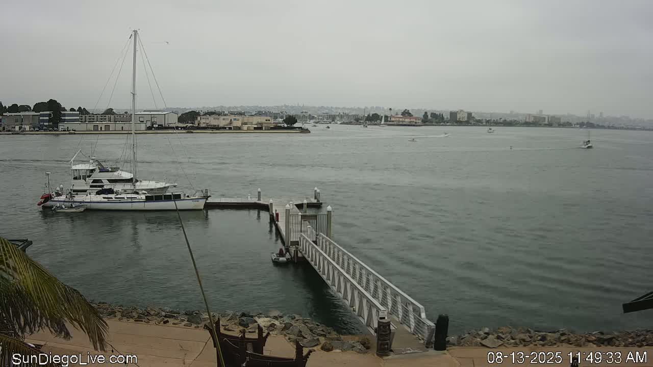 A sailboat is moored at a dock on a calm, overcast day, with other boats visible in the distance across the water.