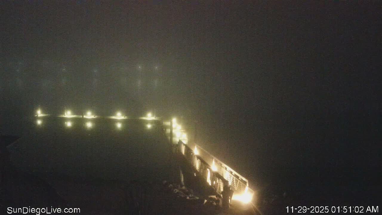 A brightly lit pier extends into dark, foggy waters at night, with its lights reflecting on the surface and disappearing into the dense haze.
