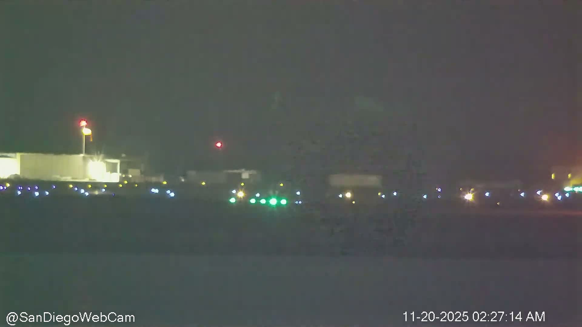 An outdoor scene at night shows an airport runway illuminated by various colored lights, with distant buildings and a windsock visible through hazy or foggy conditions.