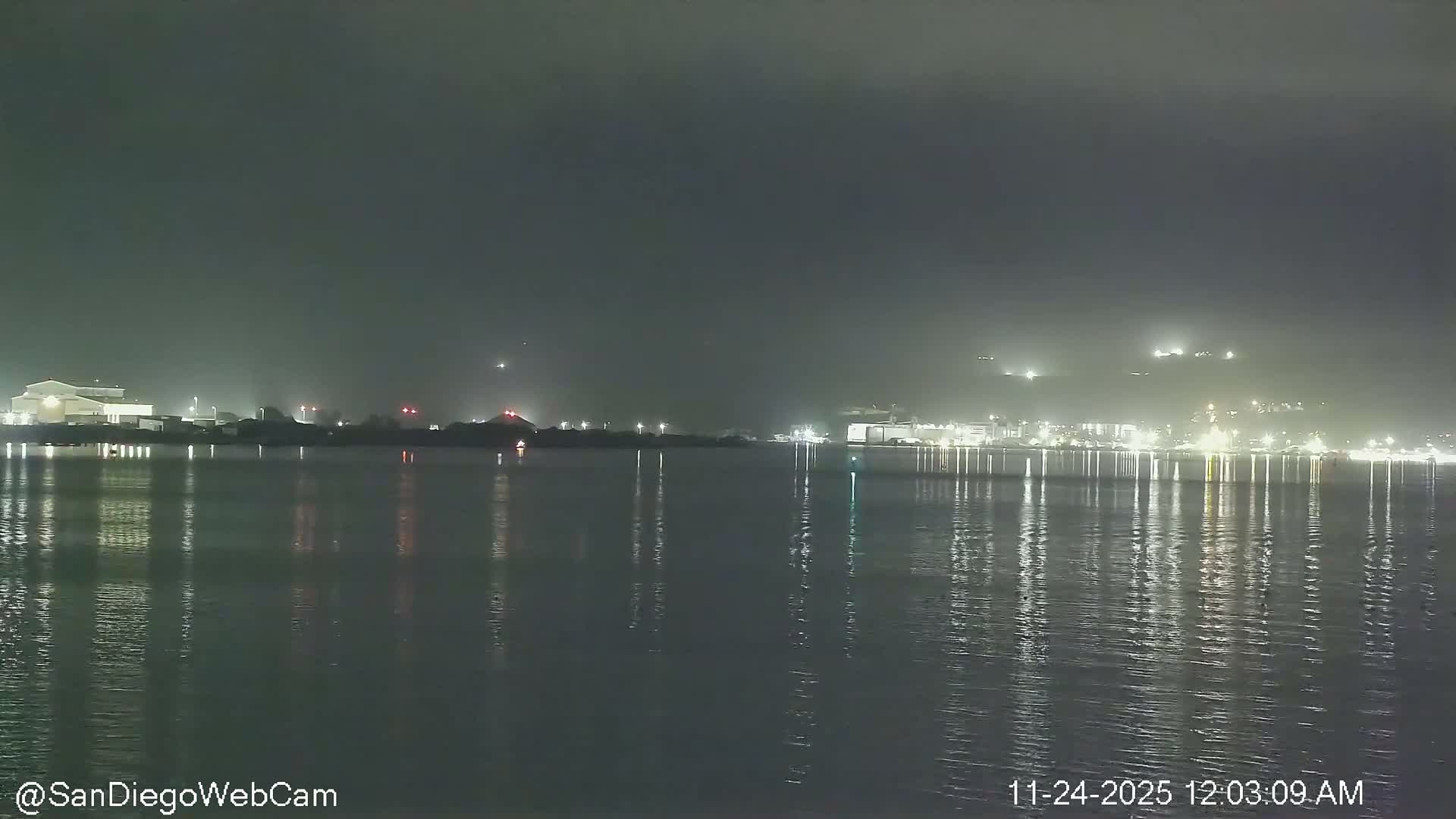 The image shows an overcast night over a body of water with the illuminated distant shoreline reflecting on its calm surface.