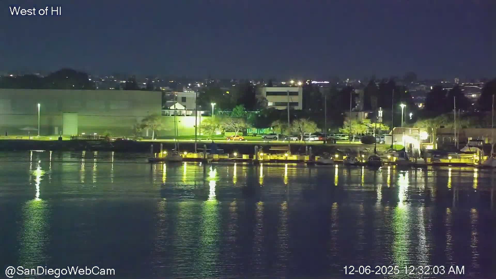 A nighttime view reveals a calm harbor filled with docked boats, reflecting the bright lights of the adjacent city and its buildings under a clear sky.