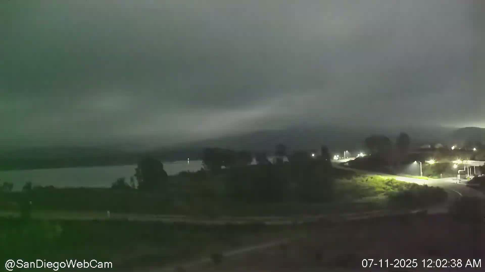 A nighttime view shows a body of water under an overcast sky, with land and vegetation in the foreground and background.