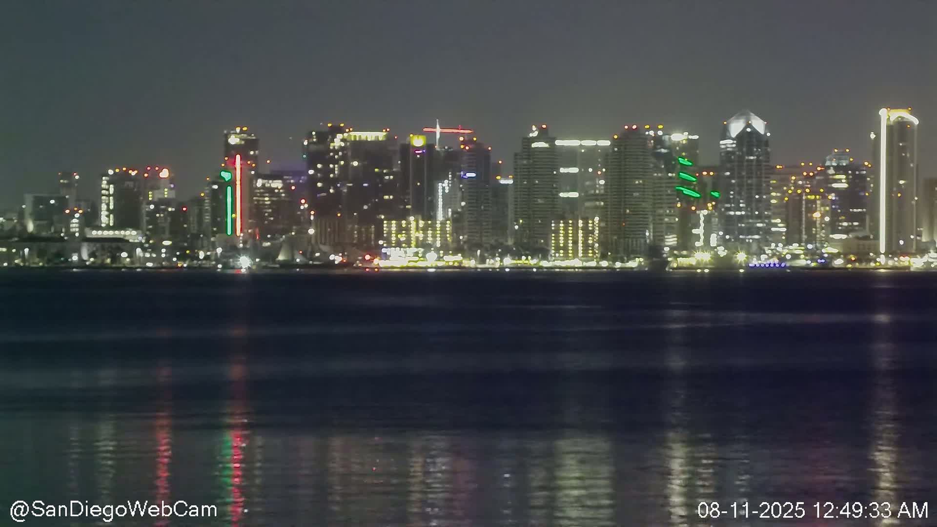 A nighttime view of a city skyline across a body of dark water with gentle waves reflecting the city lights.