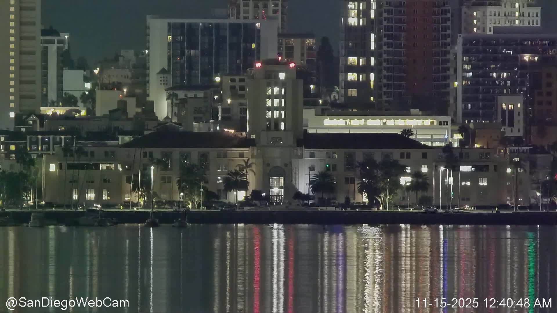A clear night view of a city skyline illuminates a calm body of water, featuring a grand central building with a prominent tower, numerous surrounding structures, and palm trees, all brightly reflected on the water's surface.