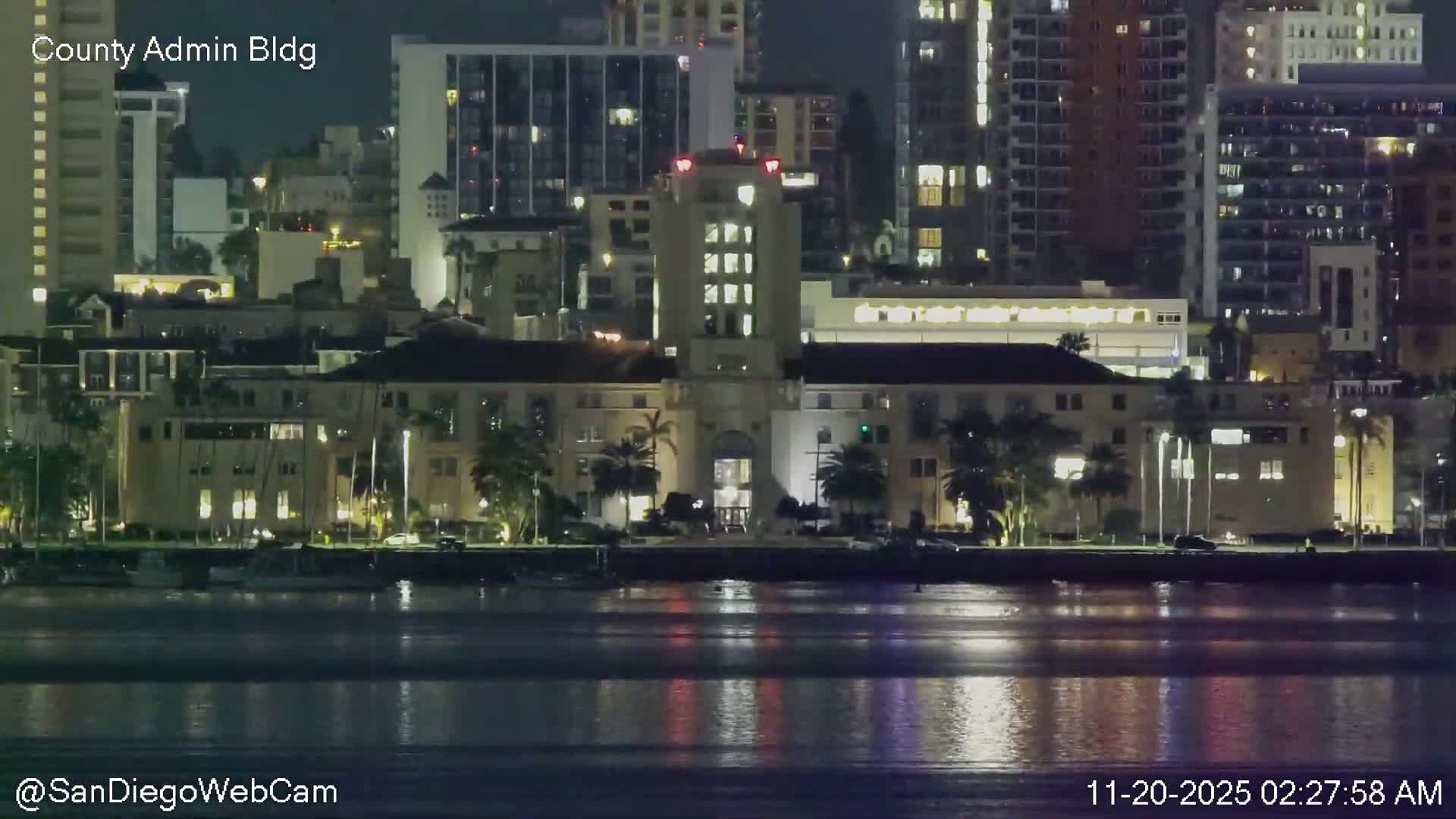 The image shows a vibrant city waterfront at night under clear skies, with brightly lit buildings, including a prominent central structure, reflecting on the calm dark water in the foreground and a dense urban skyline stretching into the background.