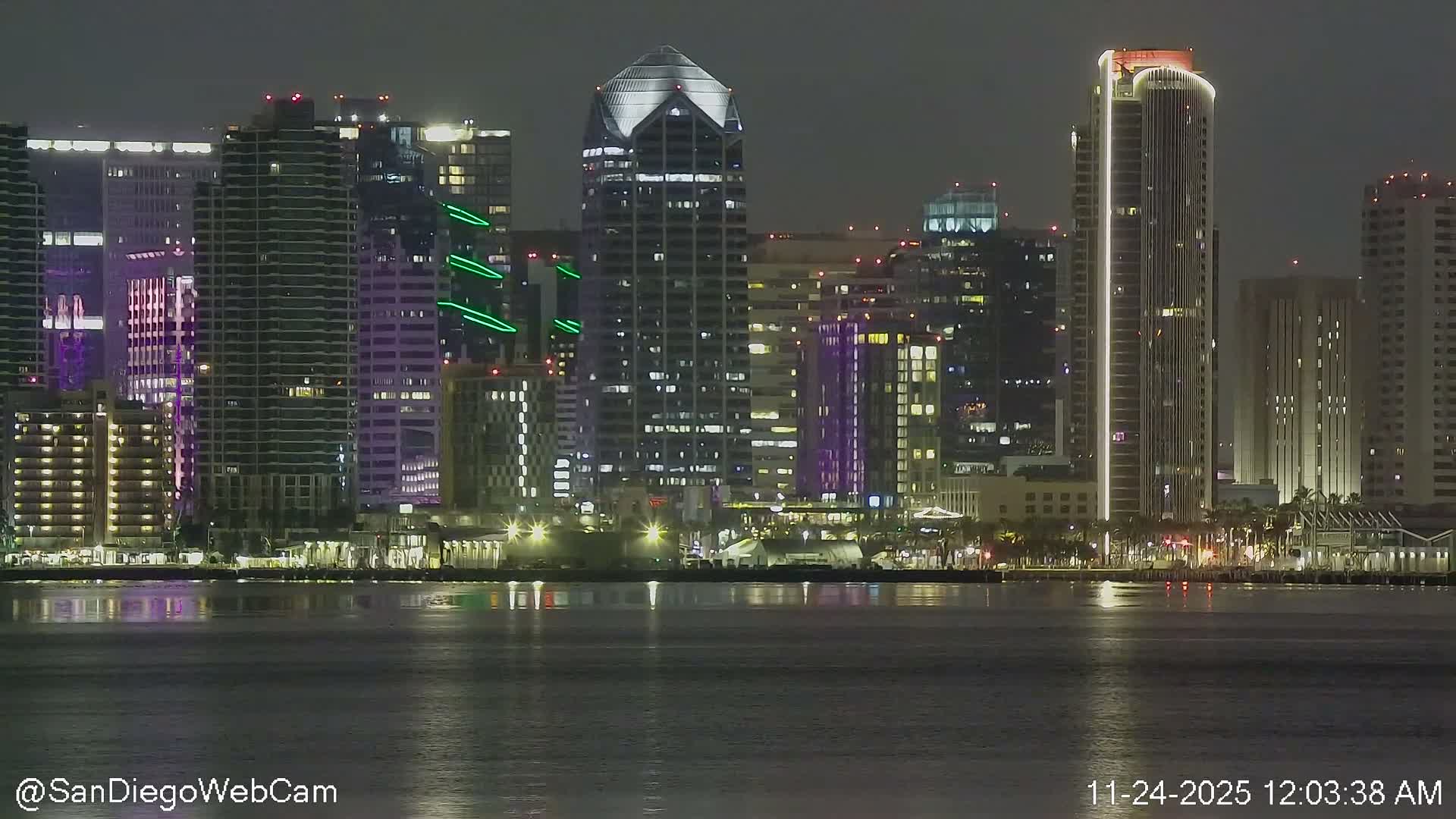 A clear nighttime scene features a brightly illuminated city skyline with numerous modern high-rise buildings, including ones with distinctive pyramidal and vertically-striped lighting designs, all reflecting on the calm dark water.
