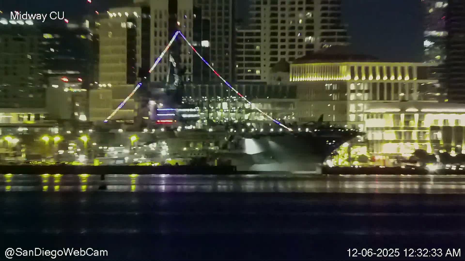 On a clear night, a large naval vessel is docked by a brightly lit city waterfront featuring a prominent statue illuminated with red, white, and blue lights, all reflected in the calm water.