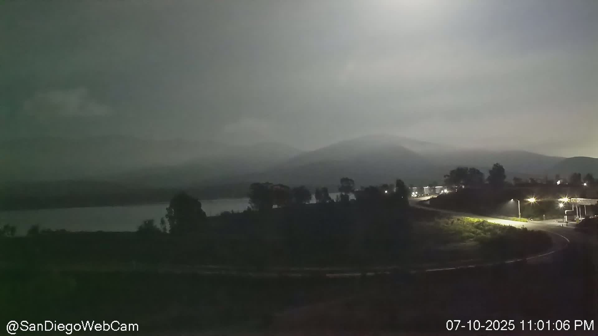 A nighttime view of a body of water, silhouetted hills, and a roadway under a hazy, moonlit sky.