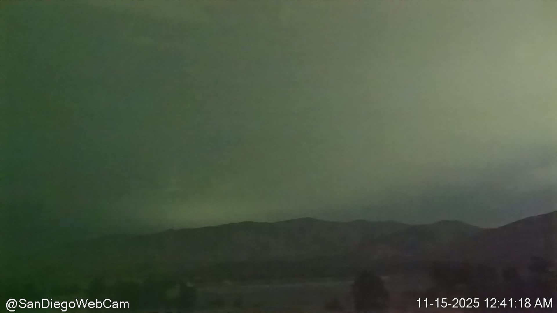 A dark, heavily overcast sky with a prominent green tint looms above a silhouetted mountain range and a shadowy foreground, indicating a very dark night or pre-dawn scene.