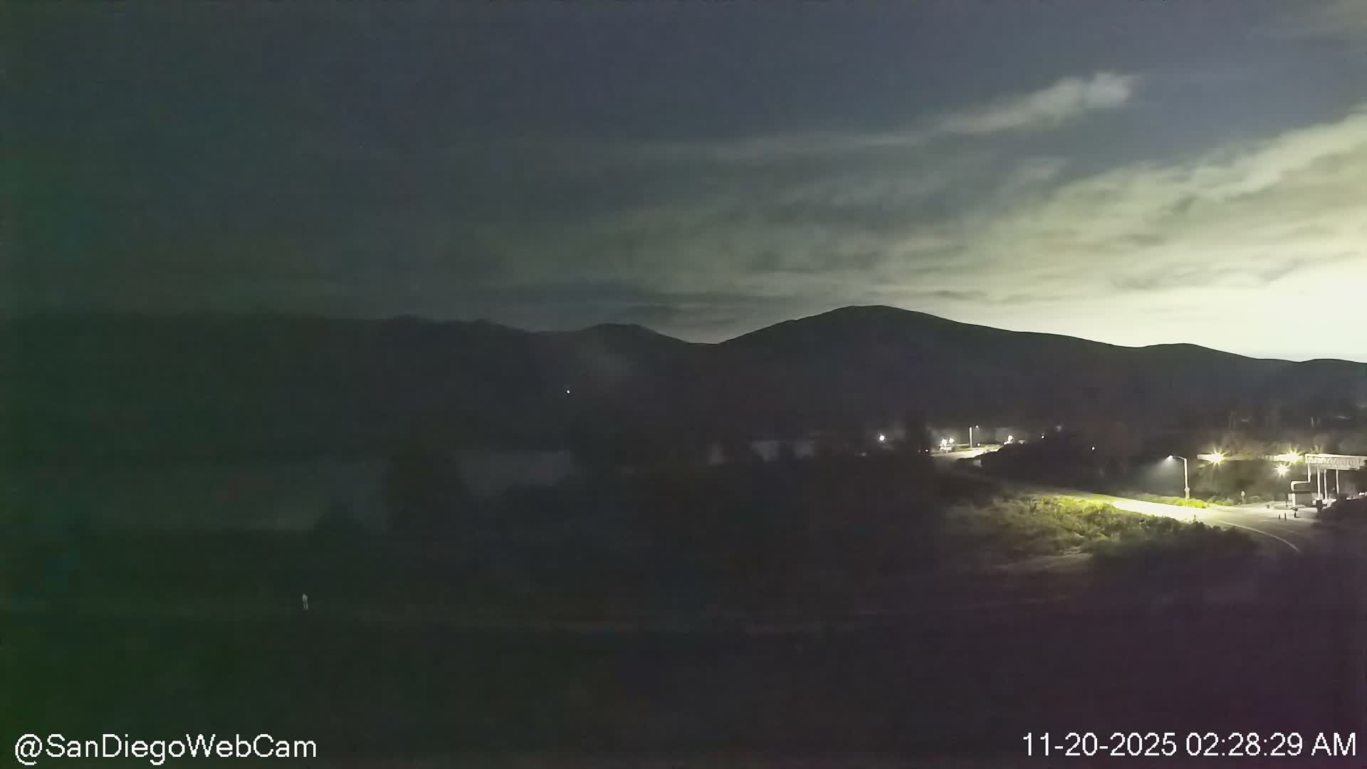 A dark, hazy night view reveals a cloudy sky above silhouetted mountains, a body of water, and an illuminated road with structures on the right.