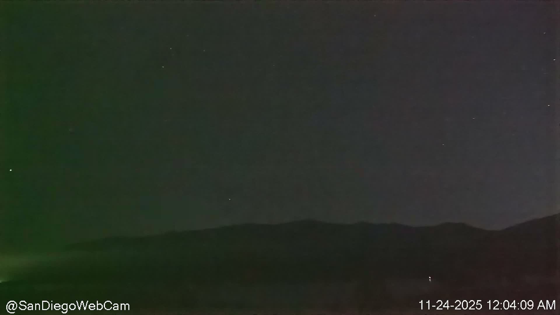 A clear, dark night sky dotted with faint stars stretches above a silhouetted mountain range, with a subtle greenish glow emanating from the left side of the distant horizon.