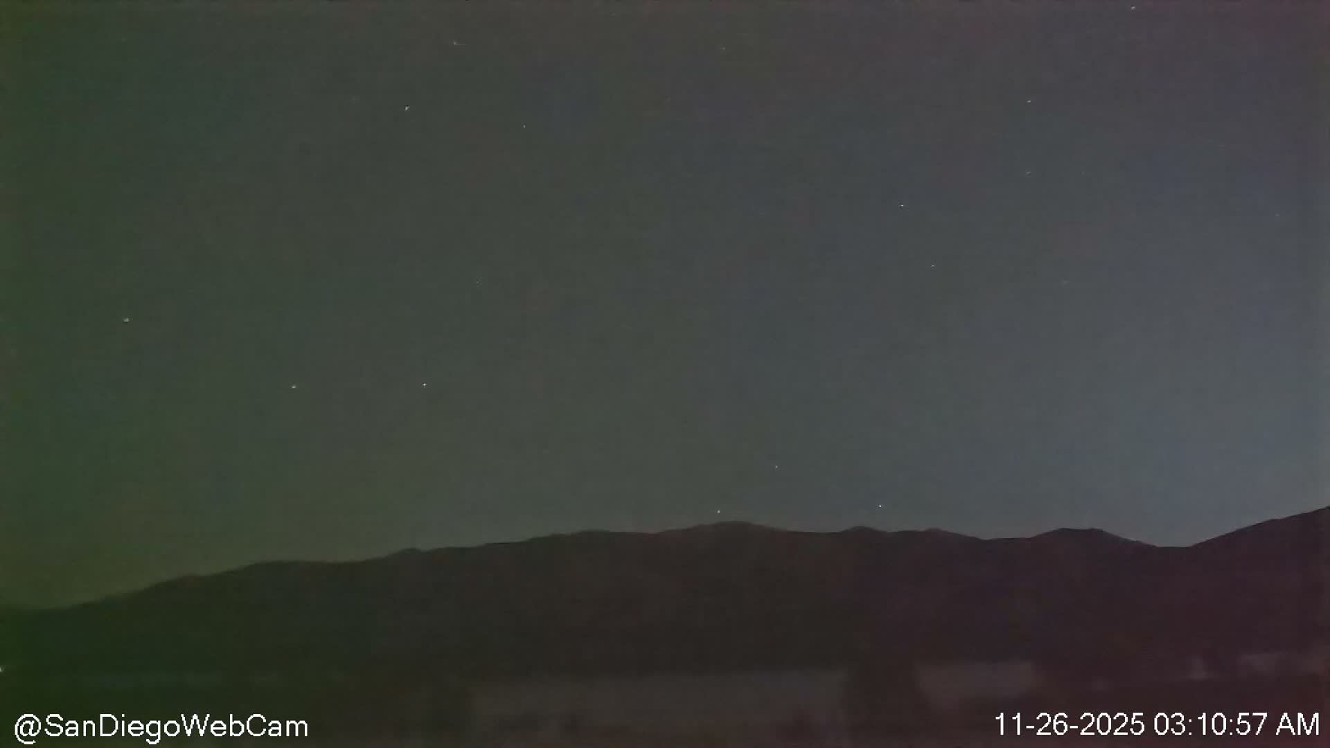A clear, dark night sky dotted with faint stars stretches above a silhouetted mountain range, with a subtle greenish glow emanating from the left side of the distant horizon.