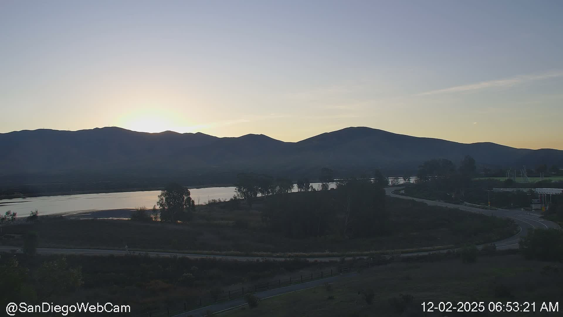 The sun rises brightly over a distant mountain range on a clear morning, casting light across a wide body of water and illuminating winding roads and trees in the foreground.