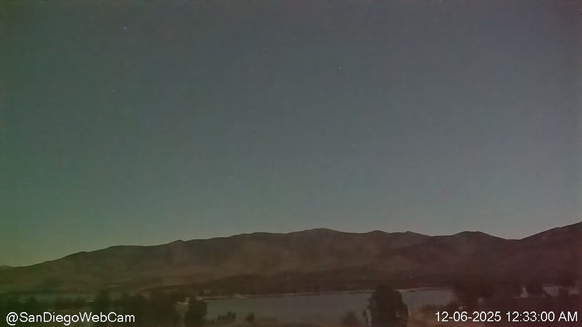 A vast, clear night sky with a few faint stars stretches over a calm body of water, flanked by dark trees and a range of distant, shadowed mountains.