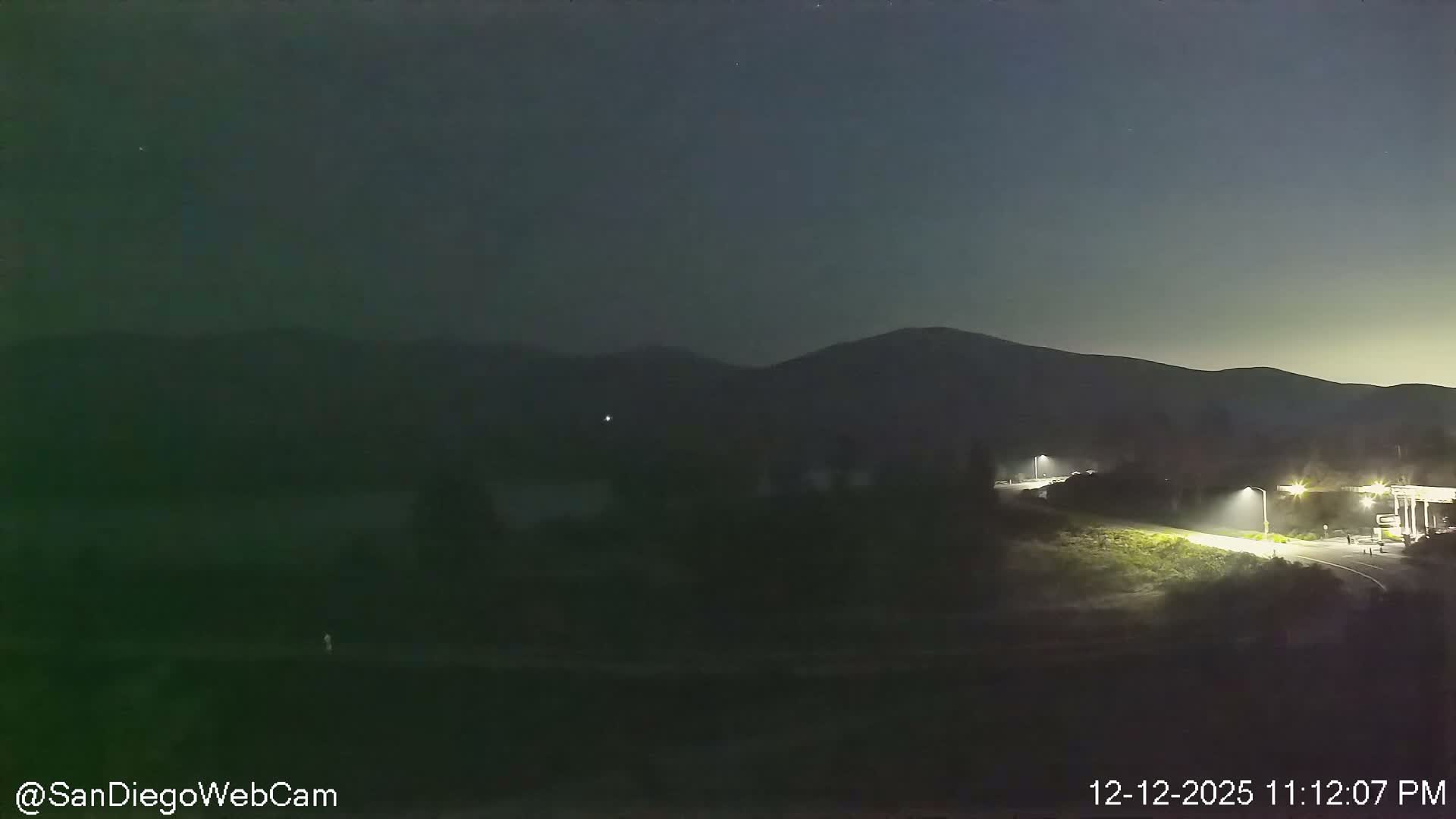 A clear night scene reveals dark mountains silhouetted against a dim sky, with a tranquil body of water in the distance and a brightly lit road leading to a gas station or service area on the right.