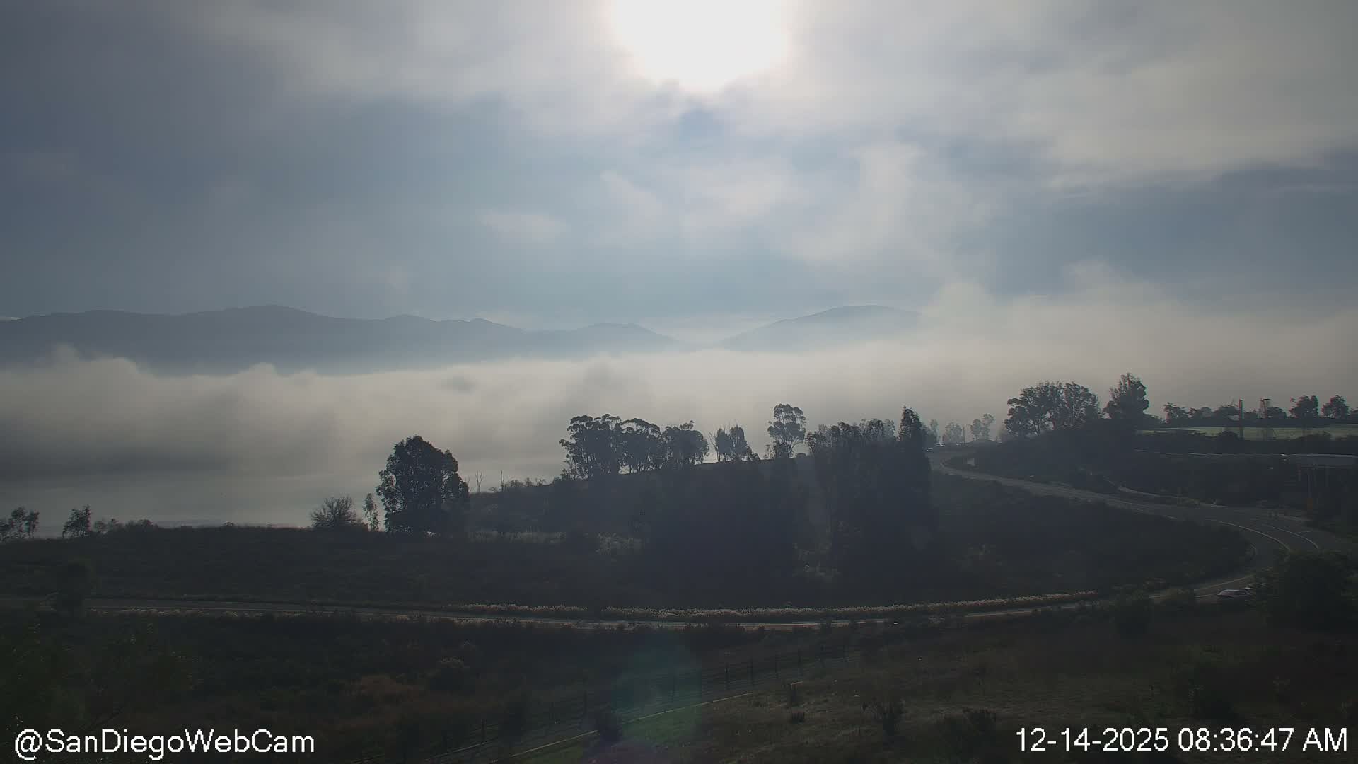 A clear night scene reveals dark mountains silhouetted against a dim sky, with a tranquil body of water in the distance and a brightly lit road leading to a gas station or service area on the right.
