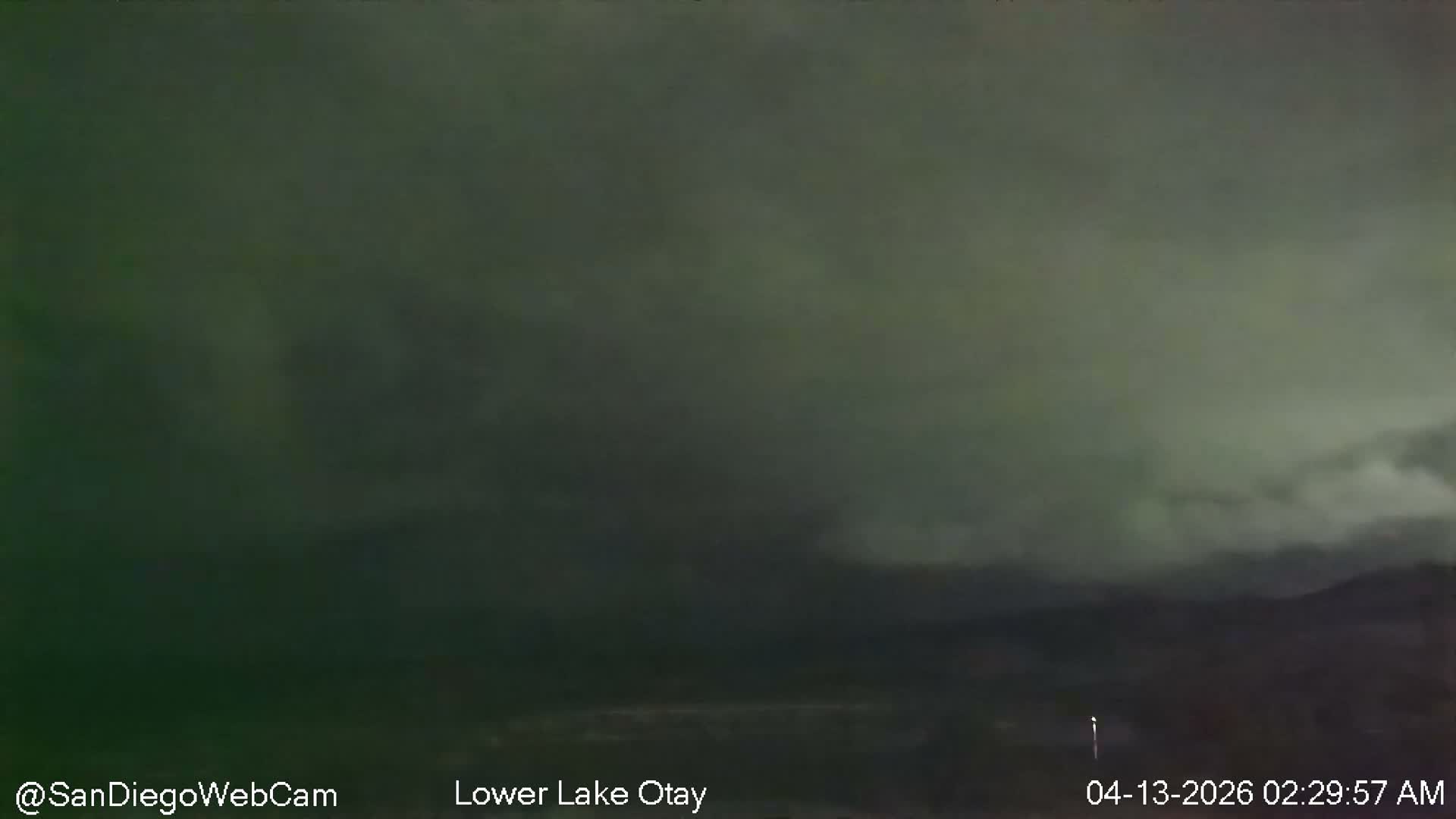 A clear night scene reveals dark mountains silhouetted against a dim sky, with a tranquil body of water in the distance and a brightly lit road leading to a gas station or service area on the right.