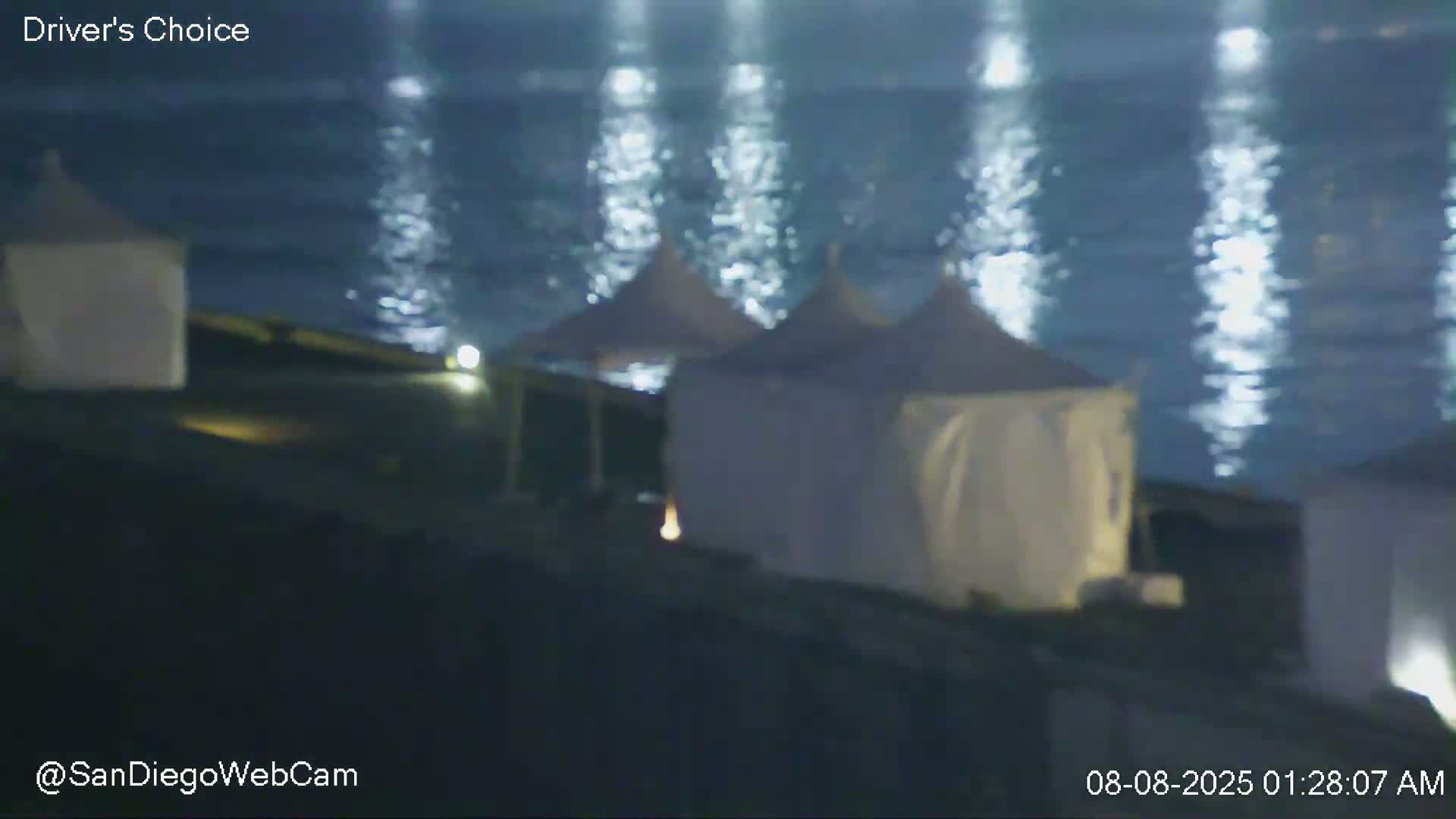 At night, several white cabanas sit on a dark surface near a body of water reflecting light.
