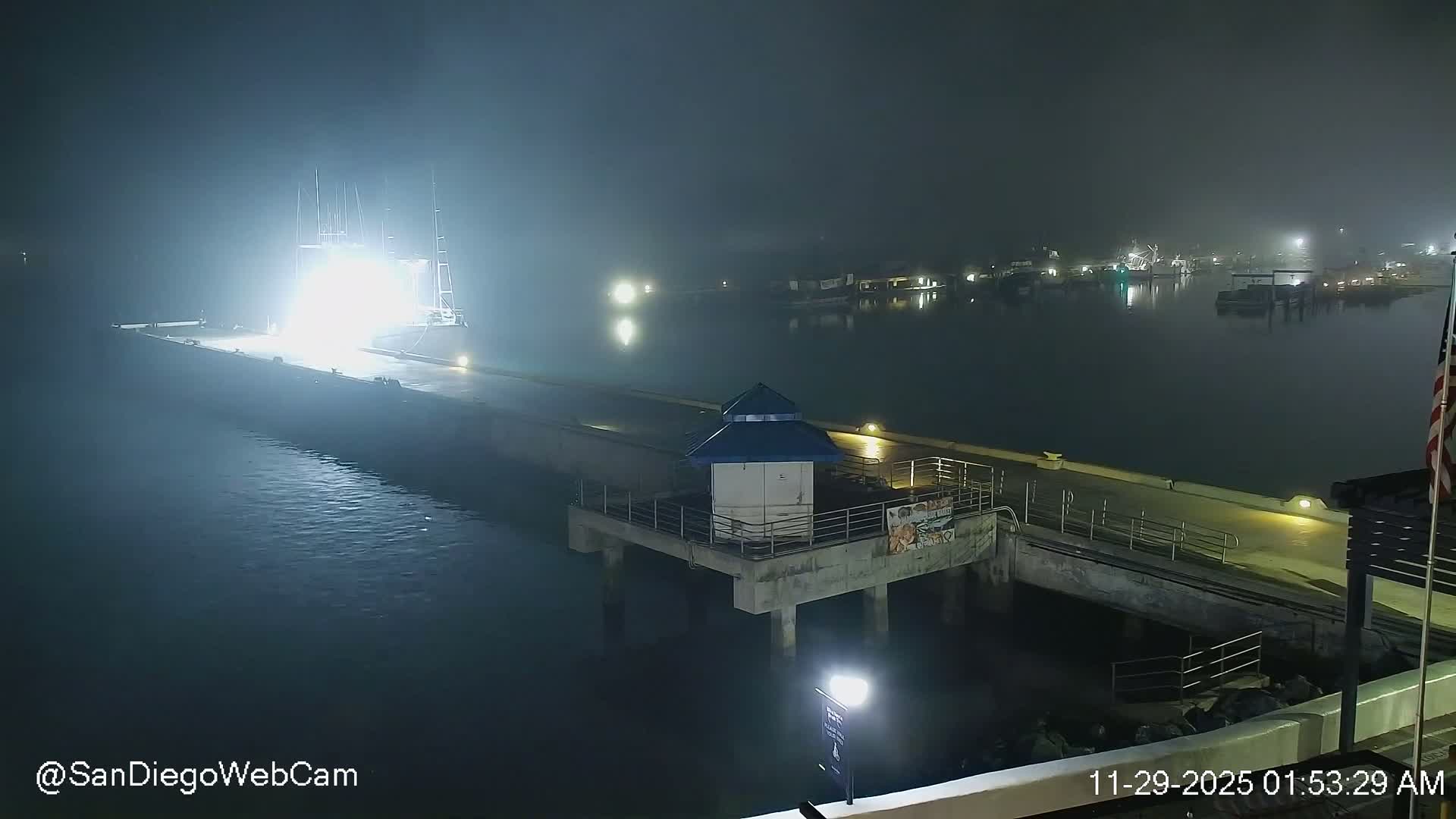 A very foggy night scene captures a brightly illuminated large vessel docked at a pier in a harbor, with a small building on a platform and distant shore lights barely visible through the thick mist.