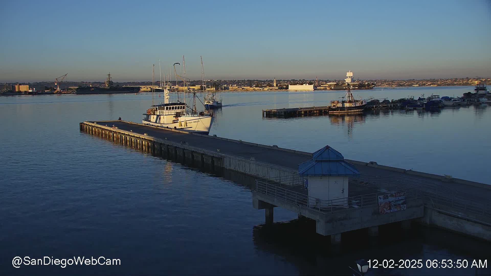 A peaceful harbor scene at early morning displays various boats docked along a long pier, with another vessel sailing further out, all beneath a clear blue sky with a distant city skyline and a large military ship on the horizon.