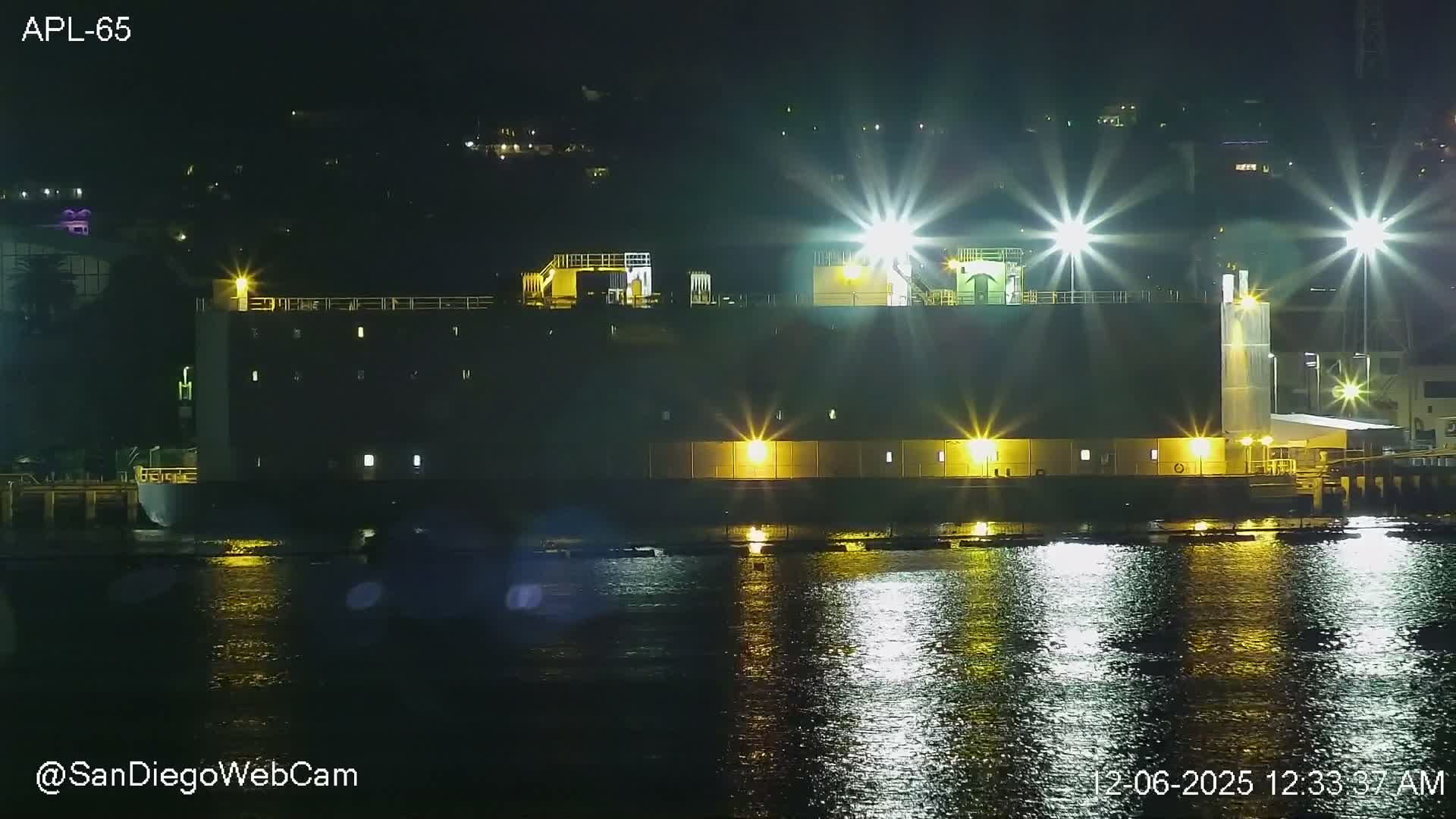 A nighttime scene displays a large, brightly illuminated industrial-like structure or ship docked next to calm water, with numerous artificial lights casting strong, star-like reflections on the dark surface under a clear sky.
