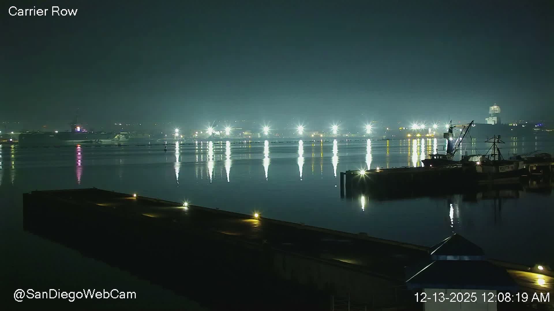 A calm harbor at night is illuminated by numerous bright shore lights reflecting on the water, with several boats docked and ships visible in the distance under a hazy sky.