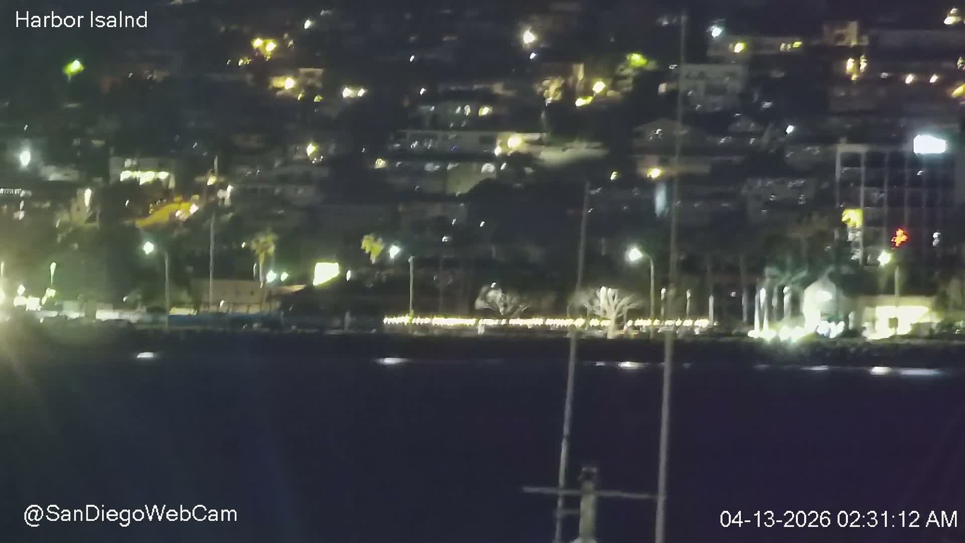A calm harbor at night is illuminated by numerous bright shore lights reflecting on the water, with several boats docked and ships visible in the distance under a hazy sky.