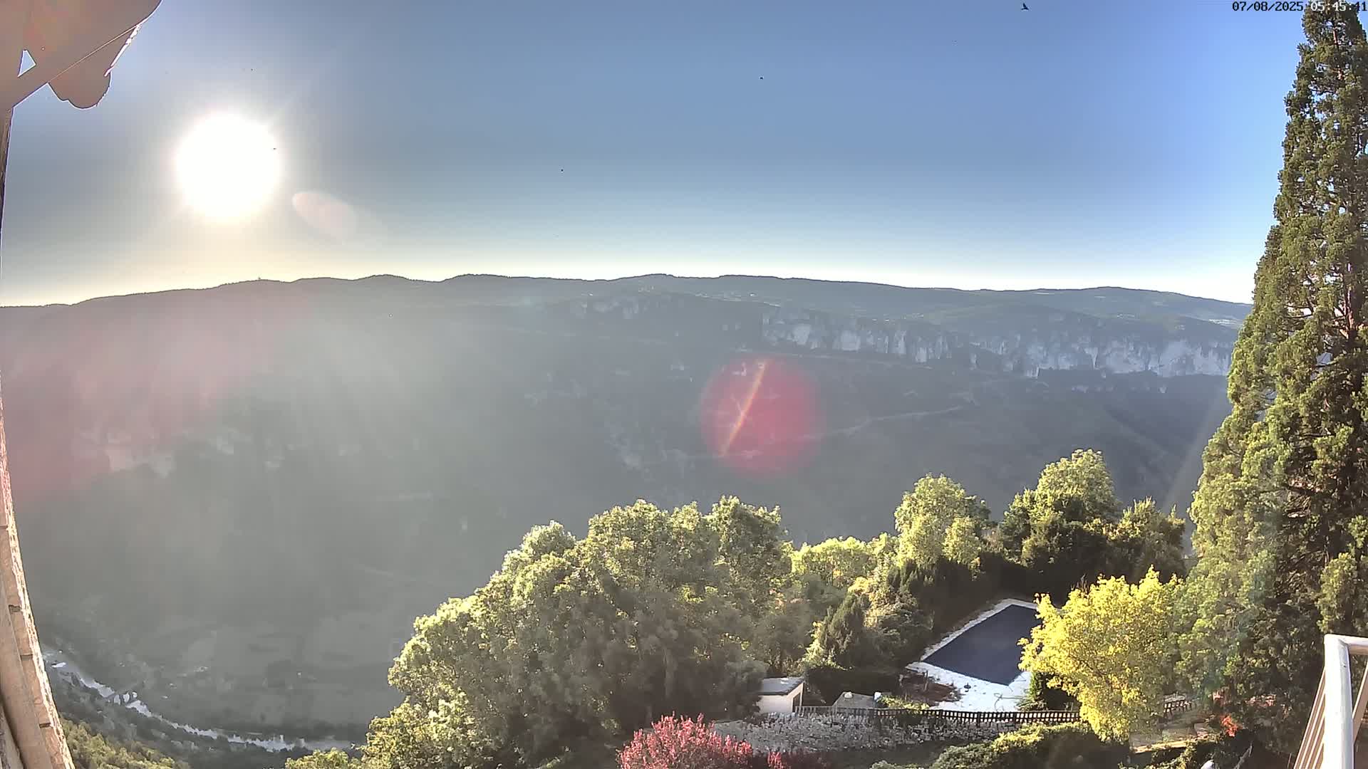 A sunny day reveals a high-angle view of a valley with a swimming pool and surrounding vegetation, overlooking a distant mountain range.