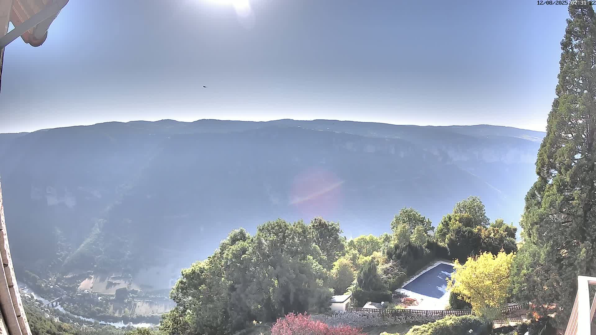 A sunny, hazy day reveals a view of a valley and distant mountains from a high vantage point, with a swimming pool and surrounding vegetation visible in the foreground.