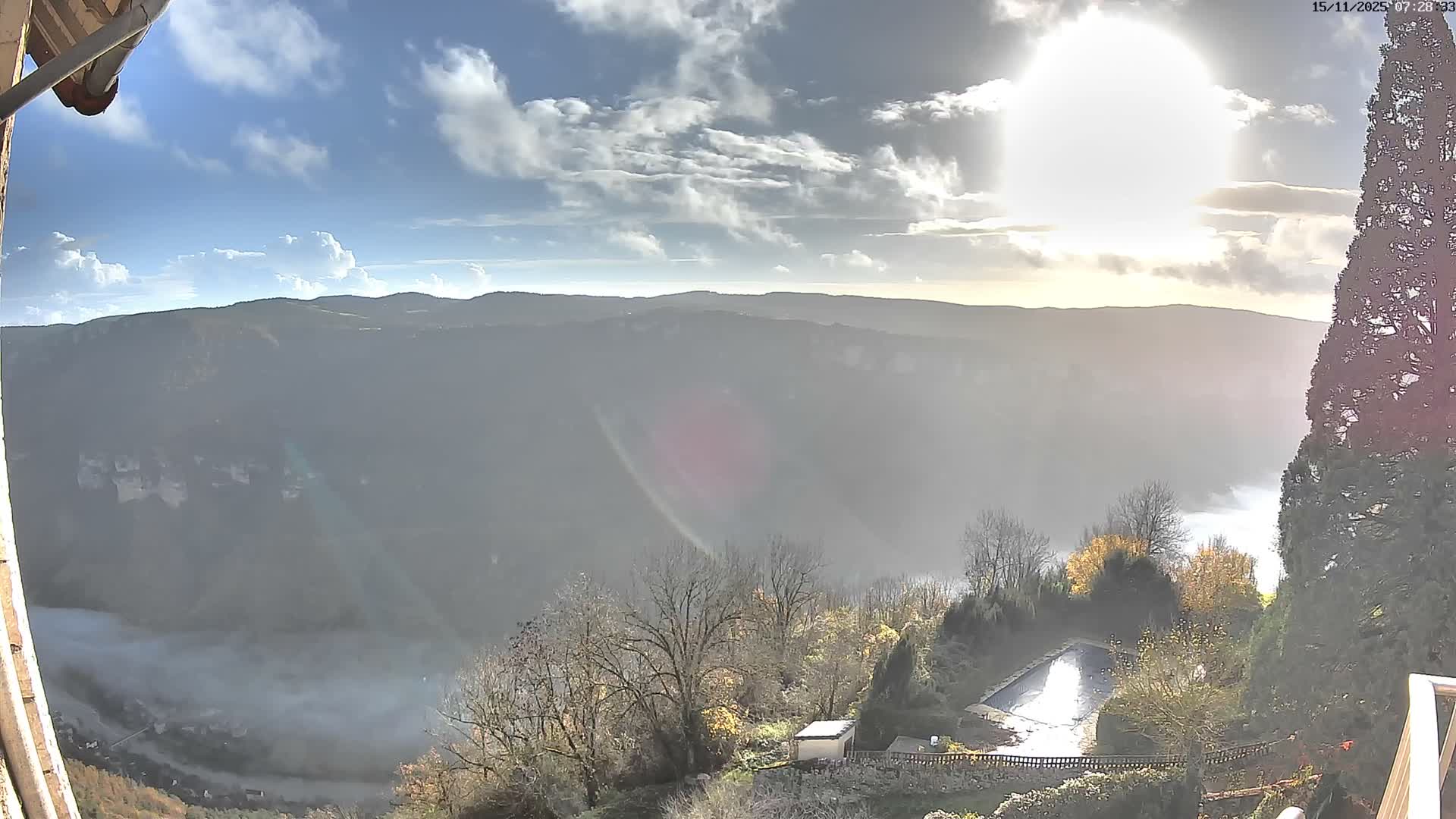 The image captures a bright, sunny morning over a wide valley partially filled with mist or fog, revealing distant mountains under a partly cloudy sky, with evergreen and bare deciduous trees framing the view from a higher vantage point.