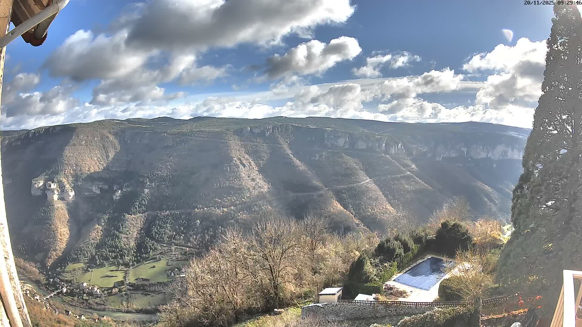 A scenic panoramic view reveals a vast, forested mountain landscape with a river and small village nestled in a valley below, framed by bare and evergreen trees in the foreground, all under a partly cloudy sky.