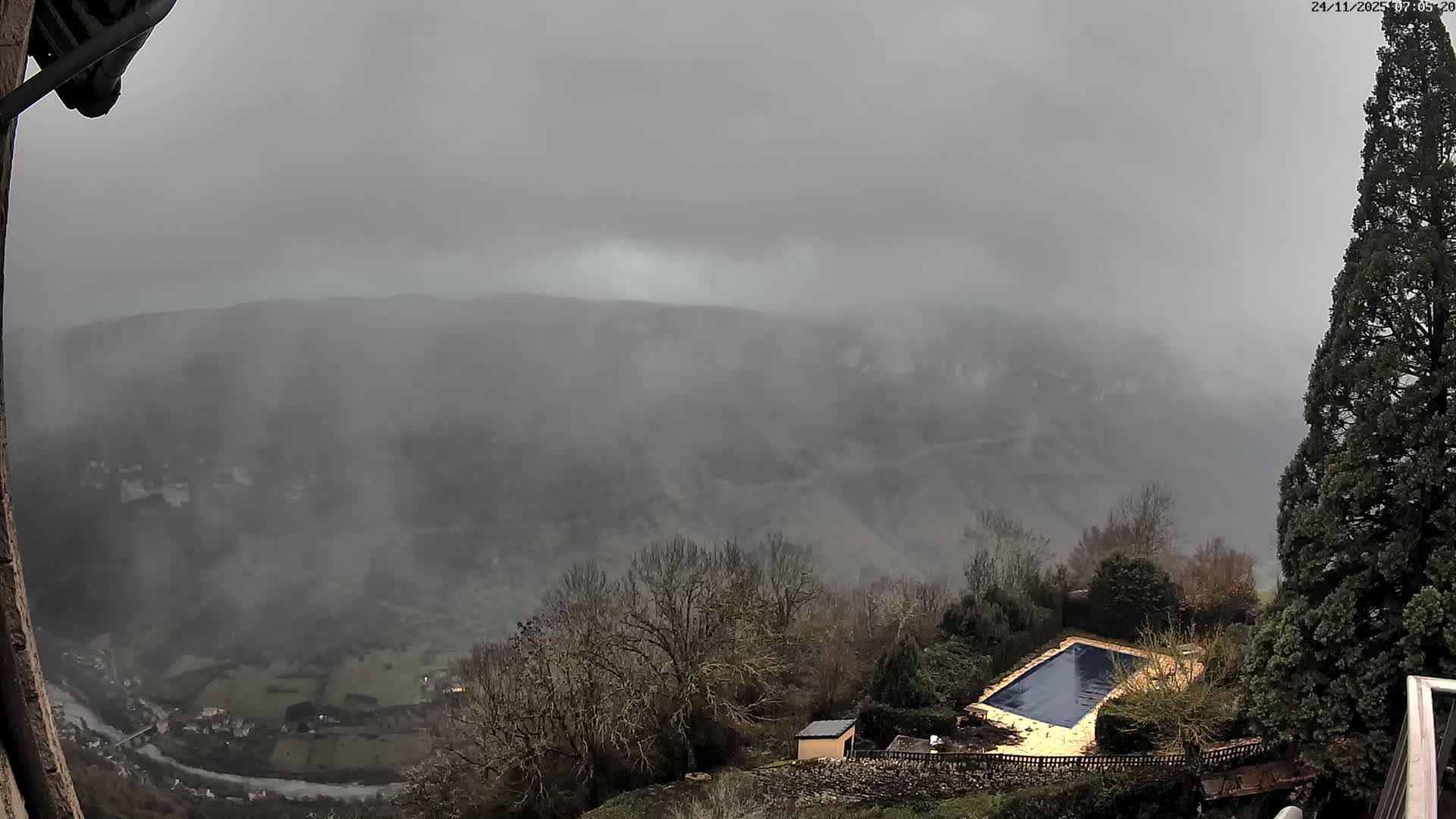 On an overcast and foggy day, a high-angle view captures a valley with a winding river and scattered buildings, framed by bare trees, a covered pool, and distant mountains shrouded in mist.