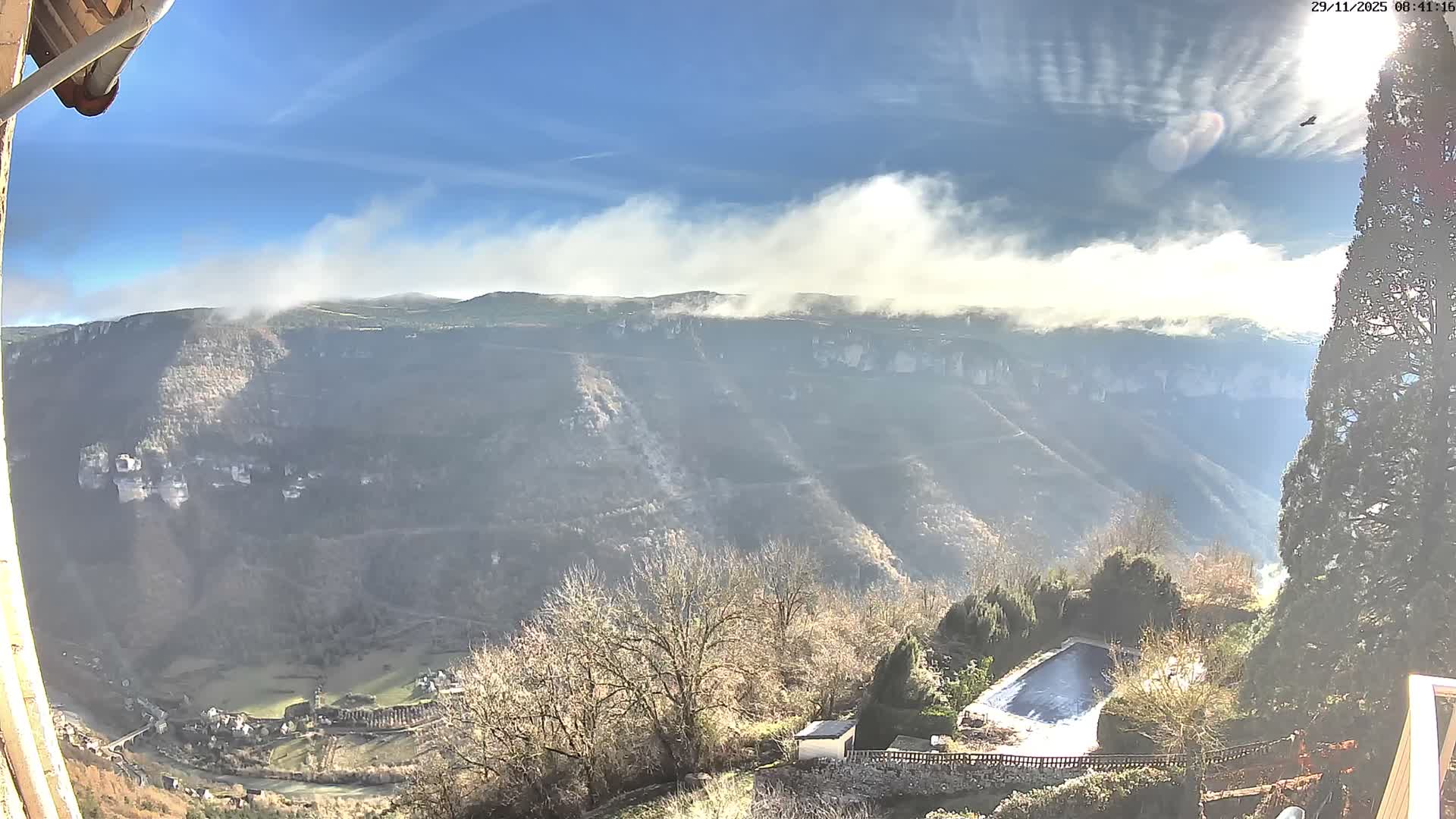 A sunny, partly cloudy panorama overlooks a deep mountain valley containing a village and river, with mist clinging to the upper ridges and a covered swimming pool among bare trees in the foreground.