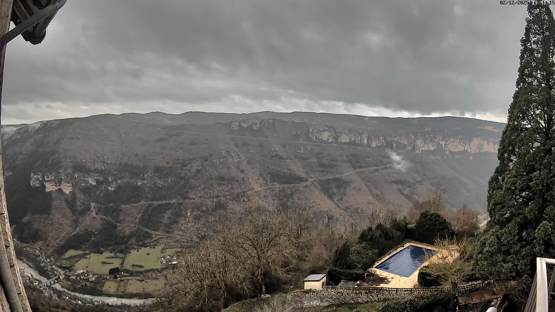 A vast valley landscape with a winding river and a small village is nestled among barren hills and distant cliffs under a heavy, overcast sky, with a covered swimming pool visible in the foreground.