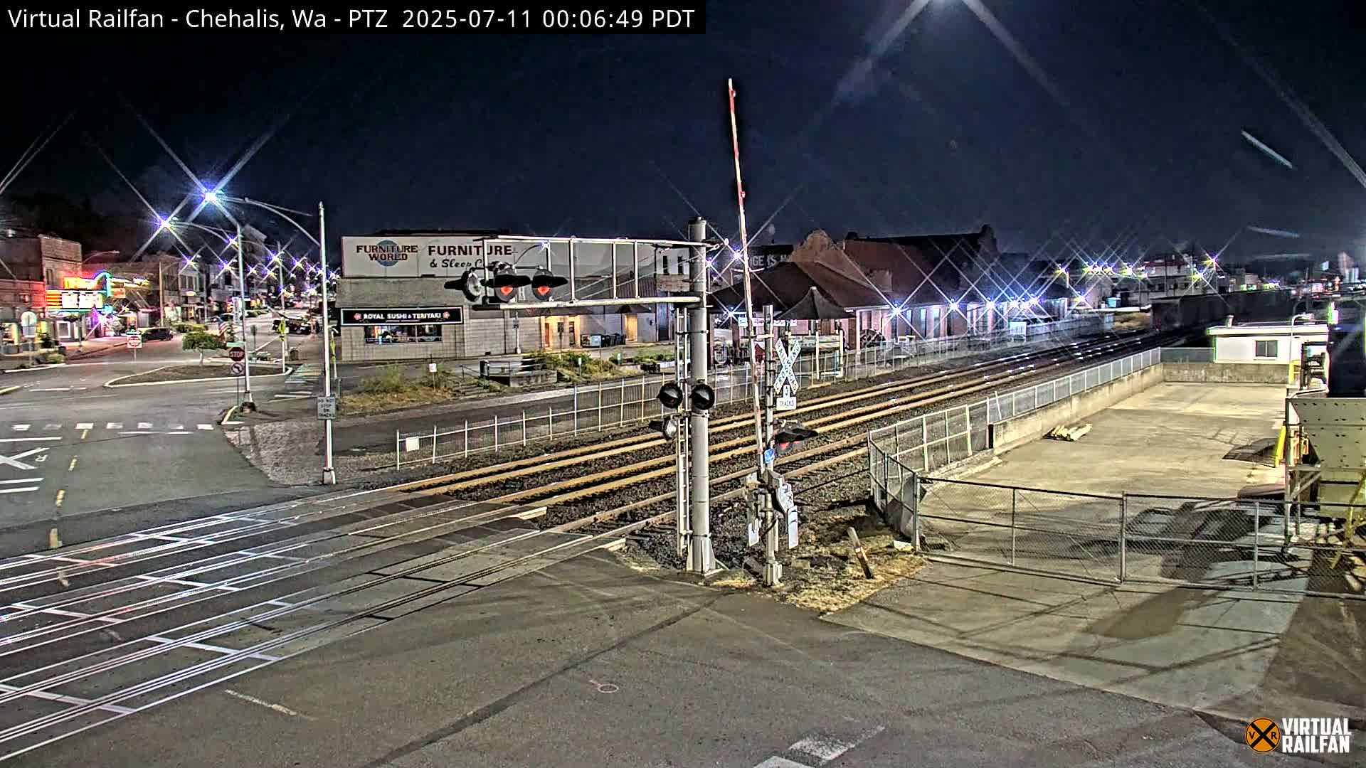A nighttime view of a railroad crossing with streetlights illuminating a mostly empty street and a train depot.