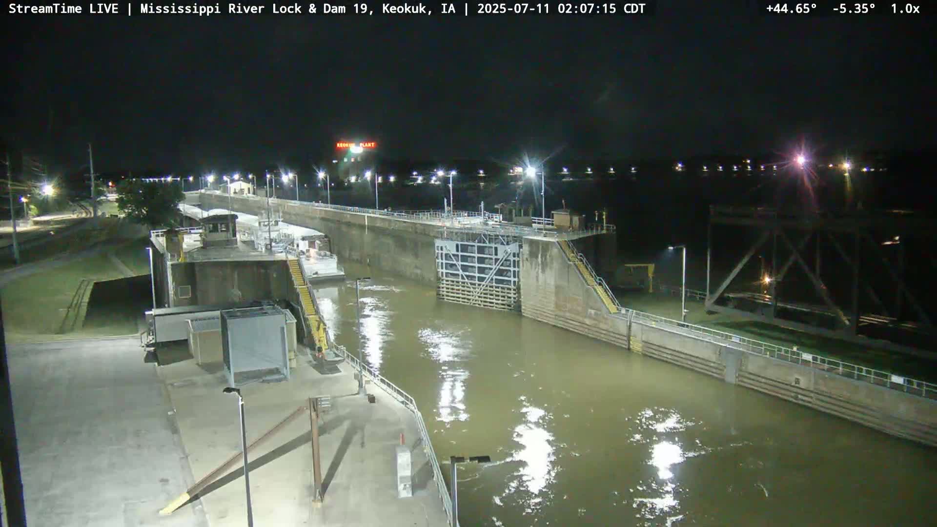 A nighttime view of a lock and dam on a river, illuminated by artificial lights.