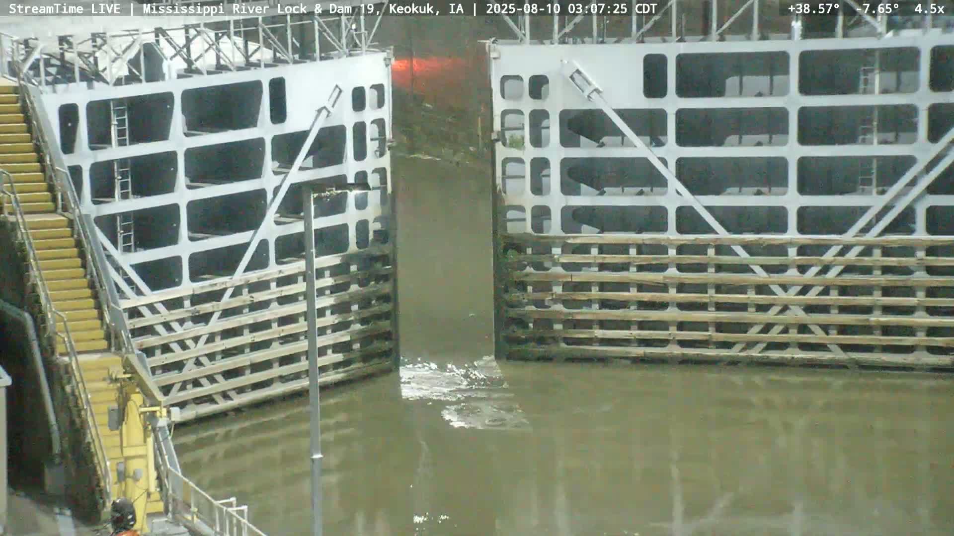 A partially opened lock gate on a river shows murky water flowing through in overcast conditions.