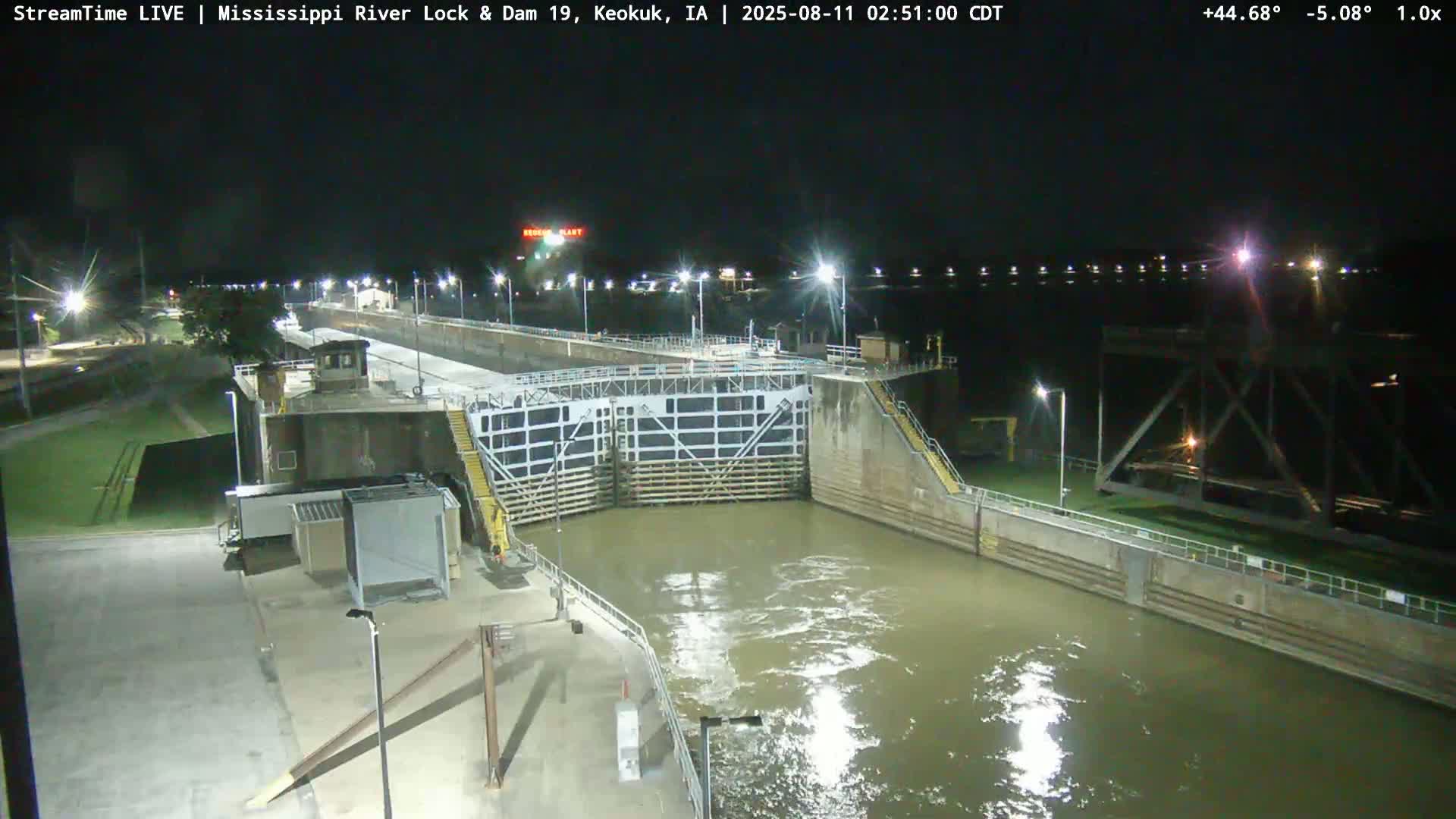 A nighttime view of a lock and dam on a river, illuminated by artificial lights.